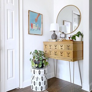 May include: A wooden cabinet with 12 drawers on hairpin legs, a round gold mirror, a white lamp with a gray base, a potted plant, and a black and white patterned stool. The cabinet has a label that reads "LEAF SUPPLY".