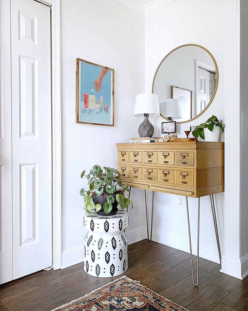 May include: A wooden cabinet with 12 drawers on hairpin legs, a round gold mirror, a white lamp with a gray base, a potted plant, and a black and white patterned stool. The cabinet has a label that reads "LEAF SUPPLY".