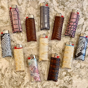 May include: A collection of 13 different patterned leather lighter cases. The cases are all in various colors and patterns, including snakeskin, crocodile, and floral.