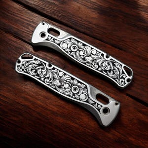 May include: Two silver knife handles with intricate floral scrollwork are displayed on a dark wood surface. The handles have a polished finish and feature cut-out details. The detailed engraving adds a touch of elegance to the design.
