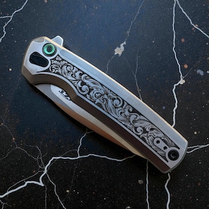 May include: A silver folding knife with an ornate floral pattern engraved on the handle. The blade is partially visible, and the knife features a green accent on the opening mechanism. The knife is set against a dark, marbled background.