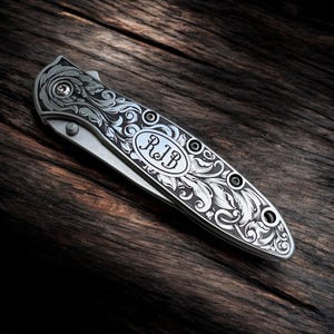 May include: A silver folding knife with intricate floral engraving. The knife features the initials "RIB" within an oval on the handle. The blade is partially open, revealing the sharp edge. The knife rests on a dark wooden surface.