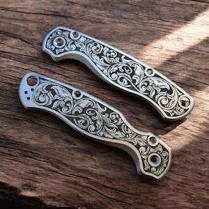 May include: Two silver pocket knives with intricate floral designs etched into the metal. The knives are laid out on a wooden surface.