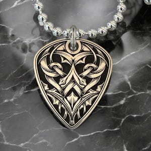 May include: A silver guitar pick necklace with a black and white intricate design. The pick is shaped like a heart and has a detailed pattern of swirls and lines.