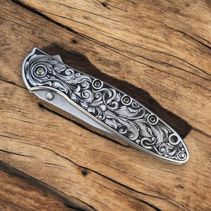 May include: A silver pocket knife with a detailed floral pattern engraved on the handle. The knife is closed and lying on a wooden surface.