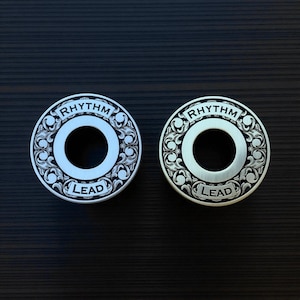May include: Two silver guitar knobs with engraved floral designs. Each knob has a central hole and the words "RHYTHM" and "LEAD" engraved on the surface. The knobs are set against a dark wood grain background.