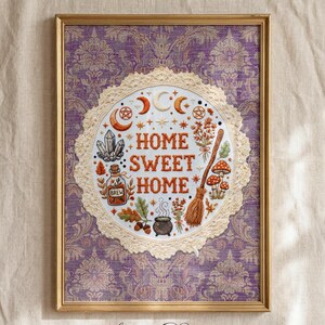 Peut inclure: Une impression d'art murale encadrée avec un fond damassé violet et un motif circulaire central bordé de dentelle crème. Le design inclut le texte "HOME SWEET HOME" entouré d'illustrations de lunes, de cristaux, d'un balai et d'un chaudron.