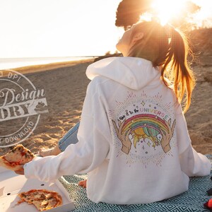 Puede incluir: Una sudadera con capucha blanca con un gr&aacute;fico de arco&iacute;ris y sol en la espalda. El gr&aacute;fico dice "Give it to the Universe". La sudadera la lleva puesta una persona sentada en una playa con una caja de pizza y una gorra de b&eacute;isbol roja.