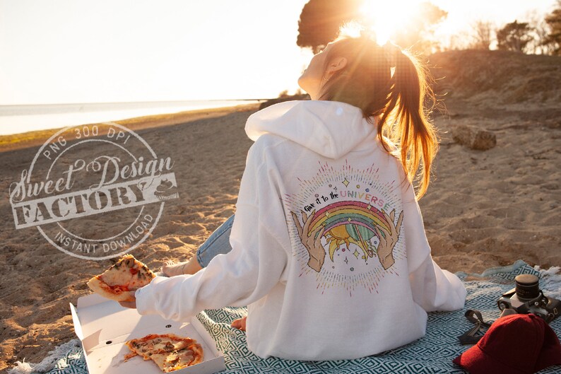 Puede incluir: Una sudadera con capucha blanca con un gr&aacute;fico de arco&iacute;ris y sol en la espalda. El gr&aacute;fico dice "Give it to the Universe". La sudadera la lleva puesta una persona sentada en una playa con una caja de pizza y una gorra de b&eacute;isbol roja.