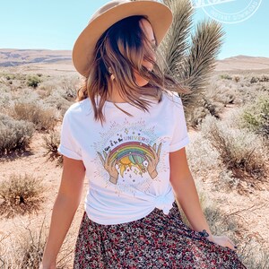 Puede incluir: Una camiseta blanca con un gr&aacute;fico de arco&iacute;ris, sol y tierra. El texto en la camiseta dice "Give it to the Universe".