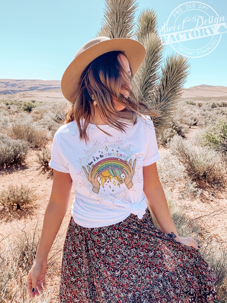 Puede incluir: Una camiseta blanca con un gr&aacute;fico de arco&iacute;ris, sol y tierra. El texto en la camiseta dice "Give it to the Universe".