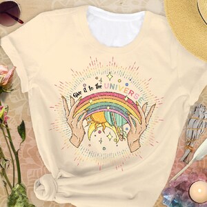Puede incluir: Una camiseta beige con un gr&aacute;fico de arco&iacute;ris, sol y planeta. El texto "Give it to the UNIVERSE" est&aacute; impreso en la camiseta.