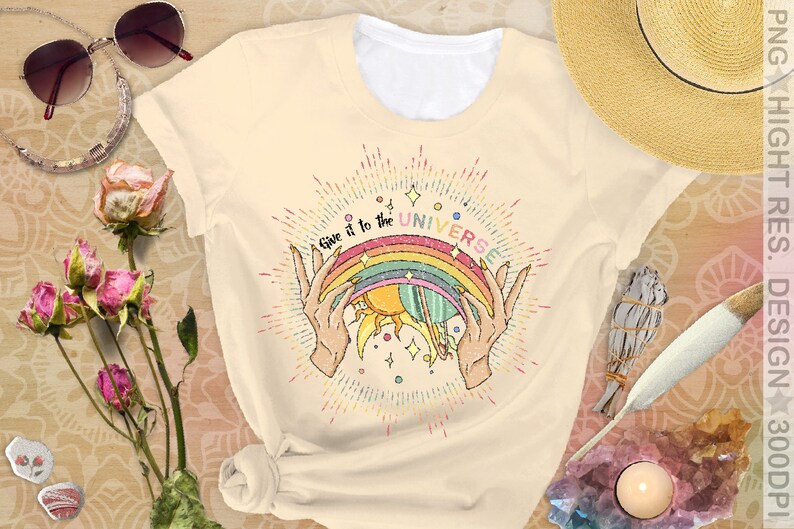 Puede incluir: Una camiseta beige con un gr&aacute;fico de arco&iacute;ris, sol y planeta. El texto "Give it to the UNIVERSE" est&aacute; impreso en la camiseta.