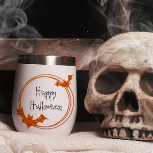 May include: White stainless steel tumbler with a gold glitter circle and the text "Happy Halloween" in black. There are two black bats in the circle. The tumbler is sitting on a white fuzzy surface next to a white skull.