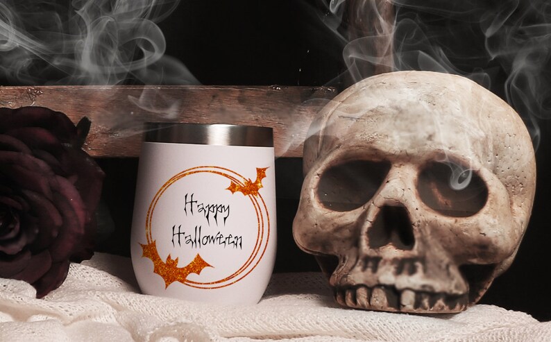 May include: White stainless steel tumbler with a gold glitter circle and the text "Happy Halloween" in black. There are two black bats in the circle. The tumbler is sitting on a white fuzzy surface next to a white skull.