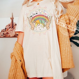 Puede incluir: Una camiseta blanca con un gr&aacute;fico de arco&iacute;ris, sol y planeta. El texto "Give it to the UNIVERSE" est&aacute; escrito en una fuente colorida.