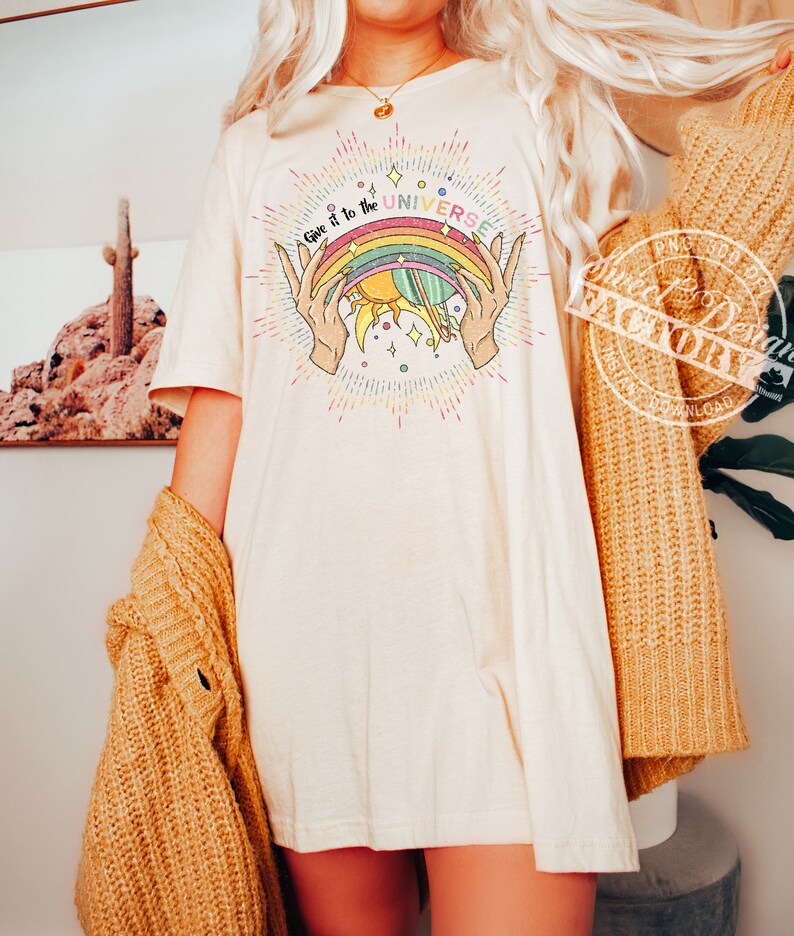 Puede incluir: Una camiseta blanca con un gr&aacute;fico de arco&iacute;ris, sol y planeta. El texto "Give it to the UNIVERSE" est&aacute; escrito en una fuente colorida.
