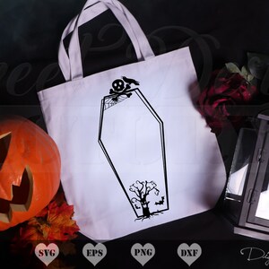 May include: A white tote bag with a black silhouette of a coffin, a skull, a spider web, and a tree. The tote bag is on a black surface with a pumpkin, fall leaves, and a black lantern.