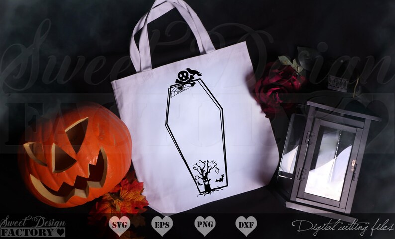 May include: A white tote bag with a black silhouette of a coffin, a skull, a spider web, and a tree. The tote bag is on a black surface with a pumpkin, fall leaves, and a black lantern.