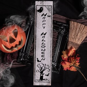 May include: A white wooden sign with the text "Happy Halloween" in black lettering. The sign features a spooky tree, bats, a ghost, and a spider web. The sign is surrounded by Halloween decorations, including a pumpkin, a broom, and fall leaves.