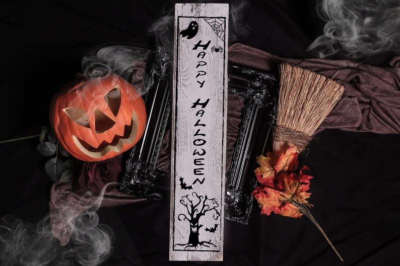 May include: A white wooden sign with the text "Happy Halloween" in black lettering. The sign features a spooky tree, bats, a ghost, and a spider web. The sign is surrounded by Halloween decorations, including a pumpkin, a broom, and fall leaves.