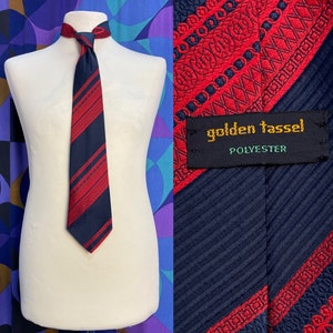 Może przedstawiać: Krawat w pasy w kolorze granatowym i czerwonym z powtarzającym się wzorem. Krawat ma metkę z napisem "golden tassel" i "polyester".