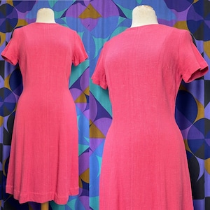 May include: A vintage, knee-length dress in a vibrant shade of pink. The dress features short sleeves, a round neckline, and vertical seam detailing. The dress is displayed on a mannequin, showcasing its tailored fit and classic silhouette.