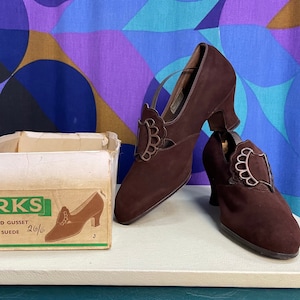 May include: A pair of brown suede Clarks Mayfield Gusset shoes with decorative silver accents, next to the original shoe box. The box features the Clarks logo and shoe details. The shoes have a small heel.