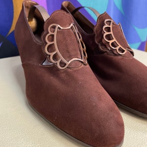 May include: A pair of brown suede shoes with decorative silver-colored accents. The shoes have a low heel and a rounded toe. The silver accents are in a scalloped design. The shoes are displayed on a cream-colored surface.