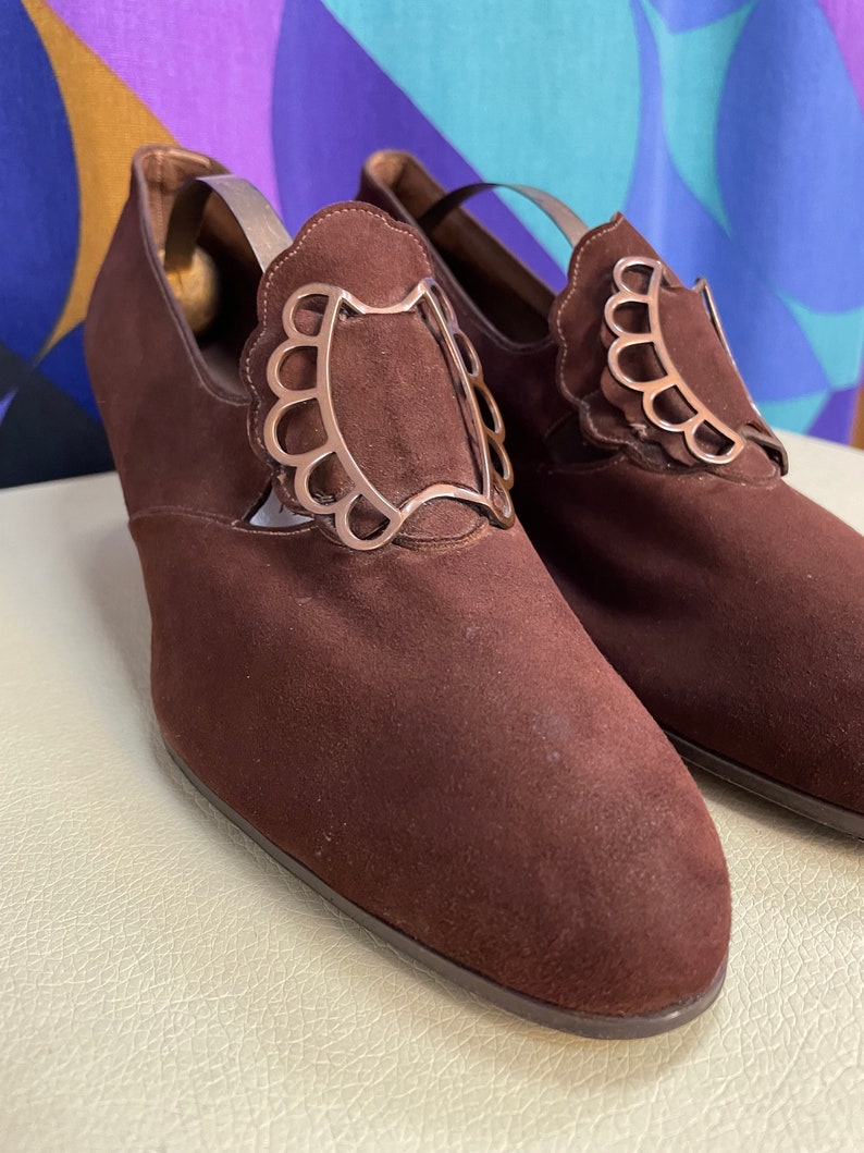 May include: A pair of brown suede shoes with decorative silver-colored accents. The shoes have a low heel and a rounded toe. The silver accents are in a scalloped design. The shoes are displayed on a cream-colored surface.