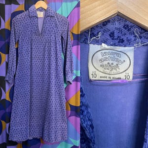 May include: A vintage purple floral dress with long sleeves and a collared neckline. The dress is displayed on a wooden hanger. The label reads "Laura Ashley" and "Made in Wales". UK size 10.