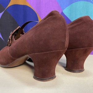 May include: Brown suede Mary Jane heels with a strap and buckle closure. The shoes have a rounded toe and a small, curved heel. The shoes are a deep brown color and appear to be vintage.