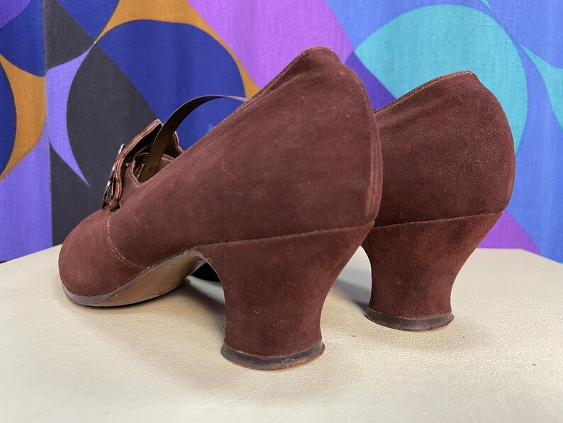 May include: Brown suede Mary Jane heels with a strap and buckle closure. The shoes have a rounded toe and a small, curved heel. The shoes are a deep brown color and appear to be vintage.