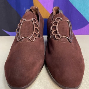 May include: A pair of brown suede shoes with decorative silver-colored metal accents. The shoes have a low heel and a pointed toe. The metal accents are in a scalloped design.