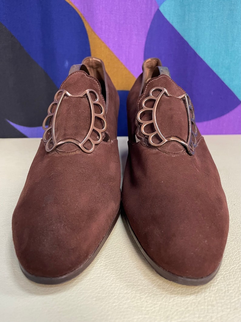 May include: A pair of brown suede shoes with decorative silver-colored metal accents. The shoes have a low heel and a pointed toe. The metal accents are in a scalloped design.