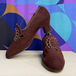 May include: A pair of brown suede vintage shoes with a low heel and decorative silver-toned metal accents. The shoes have a Mary Jane style with a strap and a scalloped design on the side.