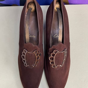 May include: A pair of brown suede Clarks shoes with decorative metal buckles. The shoes have a pointed toe and a medium heel. The shoe inserts are visible inside the shoes. The shoes are on a light green surface.