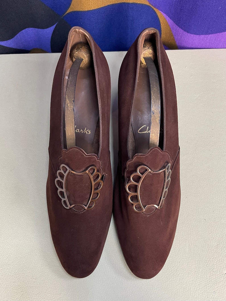 May include: A pair of brown suede Clarks shoes with decorative metal buckles. The shoes have a pointed toe and a medium heel. The shoe inserts are visible inside the shoes. The shoes are on a light green surface.