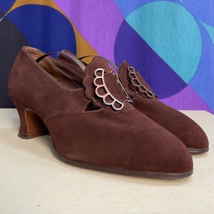 May include: A pair of brown suede vintage shoes with a decorative silver buckle. The shoes have a low heel and a strap across the top of the foot. The shoes are in good condition and appear to be unworn.