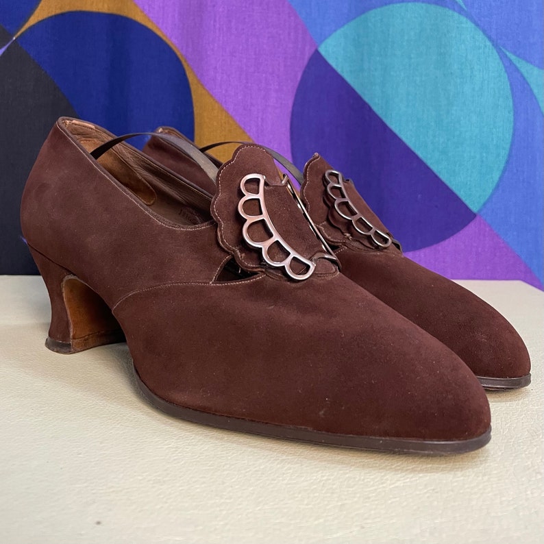 May include: A pair of brown suede vintage shoes with a decorative silver buckle. The shoes have a low heel and a strap across the top of the foot. The shoes are in good condition and appear to be unworn.