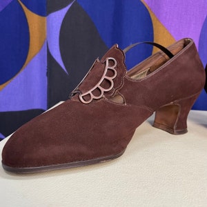May include: A brown suede shoe with a decorative silver accent. The shoe has a pointed toe, a small heel, and a strap. The silver accent is a scalloped design. The shoe is likely vintage or retro.
