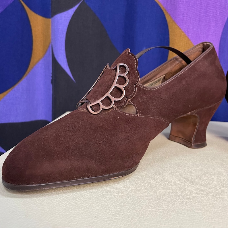 May include: A brown suede shoe with a decorative silver accent. The shoe has a pointed toe, a small heel, and a strap. The silver accent is a scalloped design. The shoe is likely vintage or retro.