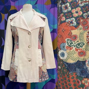 May include: A light pink corduroy coat with a notched collar and three buttons. The coat features patterned fabric panels on the front and sleeves with floral and paisley designs. The coat is displayed on a mannequin.