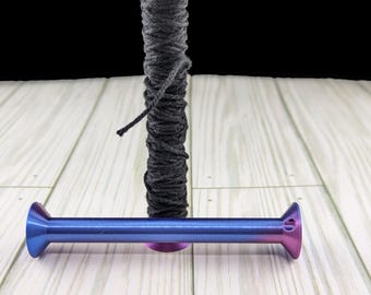 May include: Two metallic spools with a gradient finish transitioning from blue to purple. One spool stands vertically wrapped in black yarn, while the other lies horizontally in front, featuring a small hole at one end of its flared rim.