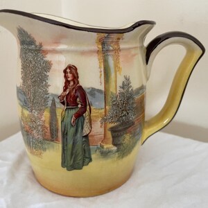 May include: A vintage ceramic jug with a landscape scene featuring a woman in a long dress. The jug has a yellow base, a brown handle, and a dark brown rim. The artwork depicts a woman standing near a column and trees.