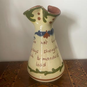 May include: A cream-coloured ceramic vase with a ruffled rim, featuring green leaf patterns, blue accents, and brown hand-painted text that reads: Do not burden todays strength with to-morrows load. The base has a thin brown horizontal stripe.