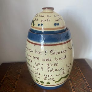 May include: A cream-colored ceramic tobacco jar with a matching lid, featuring blue and brown horizontal bands. The jar is decorated with hand-painted green accents and black handwritten text that reads: Tobacco hie! If you are well it will make you sick, Tobacco hie! It will make you well if you are sick.