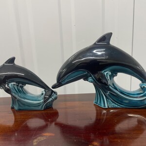 May include: Two glossy ceramic dolphin figurines in varying sizes, each featuring a dark black upper body and a teal blue wave base. The dolphins are depicted in a leaping motion, with smooth, reflective surfaces and detailed sculpted fins.