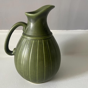 May include: Green ceramic pitcher with a handle and a spout. The pitcher has vertical ribbed detailing on the body and a rounded base. The pitcher is a solid olive green color.