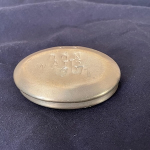 May include: A small, oval-shaped brass container with a smooth, metallic finish. The top surface features stamped lettering that reads "7 Q N A T S" and "W 10 071". The object has a rounded, low-profile design with a visible seam around the centre.